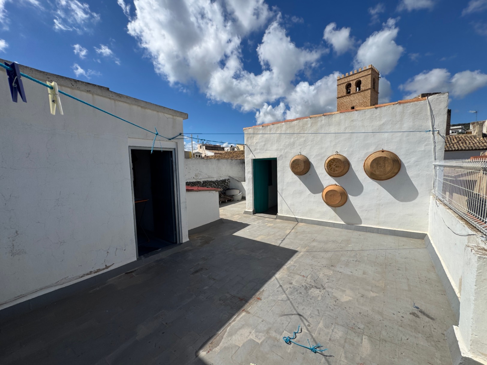 Rooftop terrace with stone tile flooring, whitewashed walls decorated with terracotta bowls, and views of a historic stone tower under a bright sky.