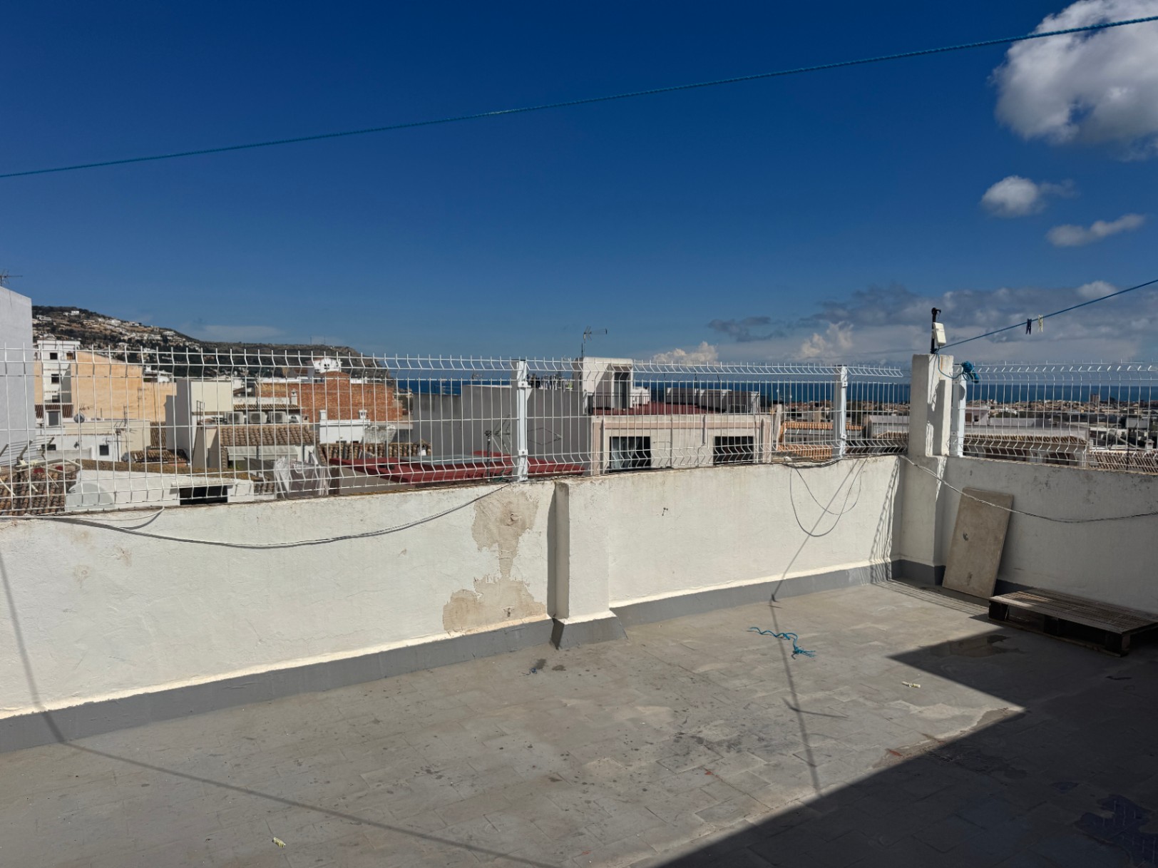 Rooftop terrace with tiled flooring and white perimeter walls topped with wire fencing, offering views of the town, distant hills, and the sea.