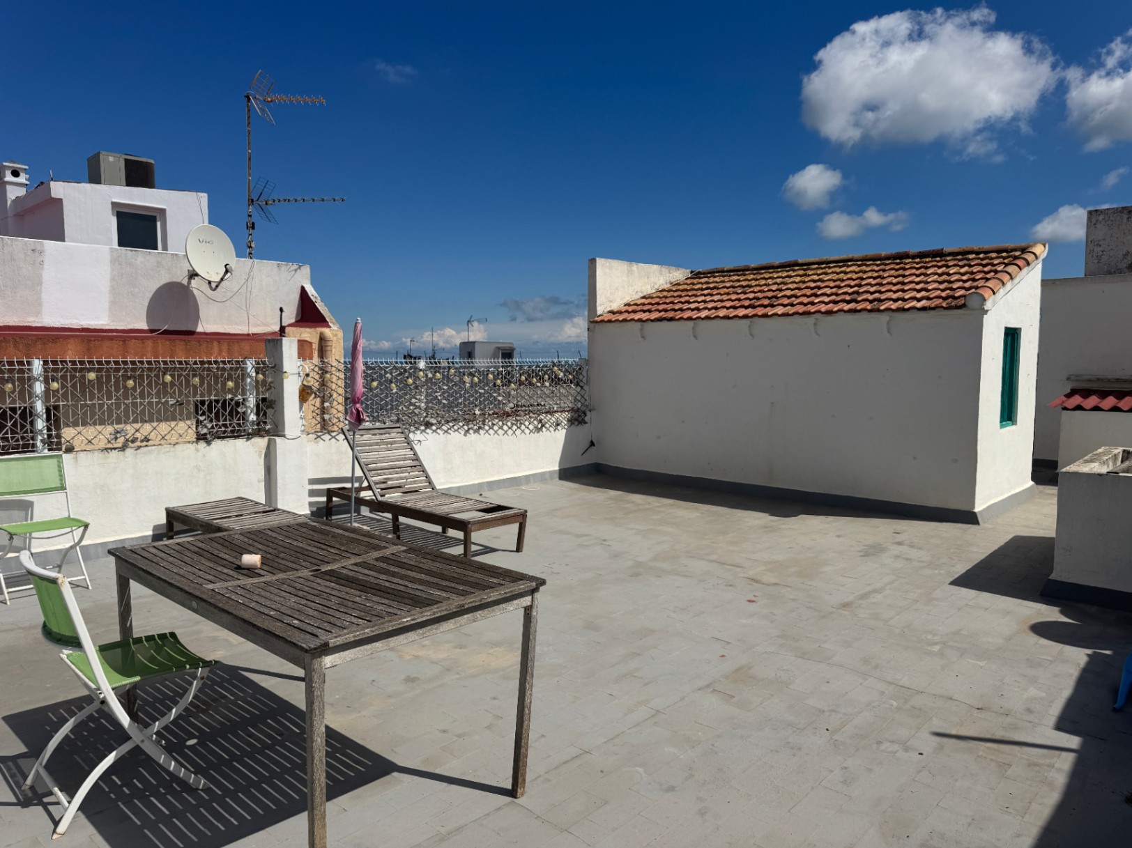 Spacious rooftop terrace with grey tile flooring, weathered wooden furniture, and white perimeter walls under a bright blue sky.