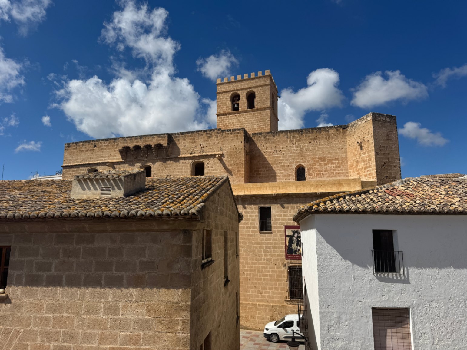 View of a historic fortified stone church with a crenellated bell tower, surrounded by traditional Mediterranean houses with clay tile roofs under a blue sky.