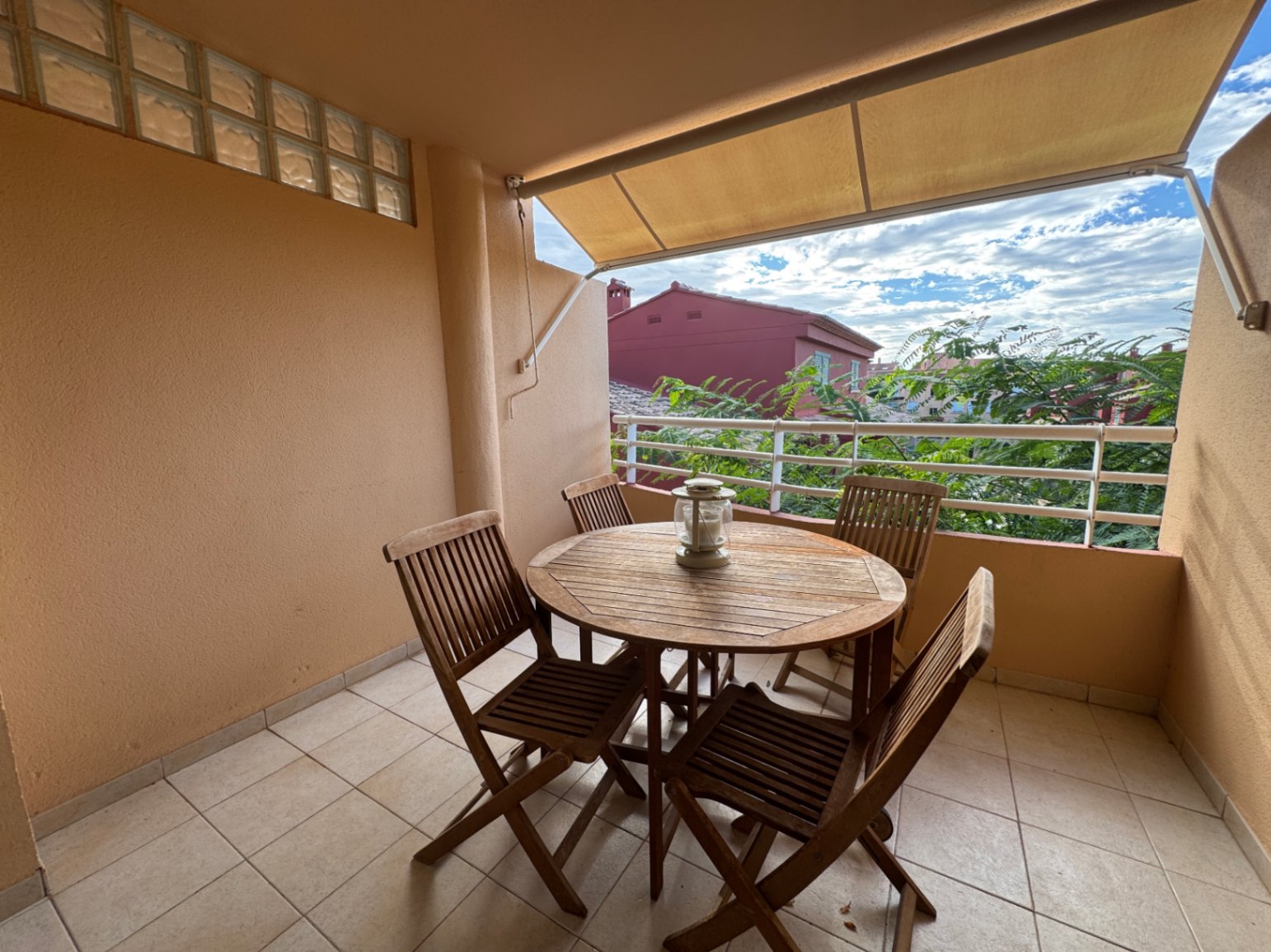 Covered terrace featuring a round wooden dining set, tiled flooring, and a retractable awning overlooking local greenery and residential buildings.