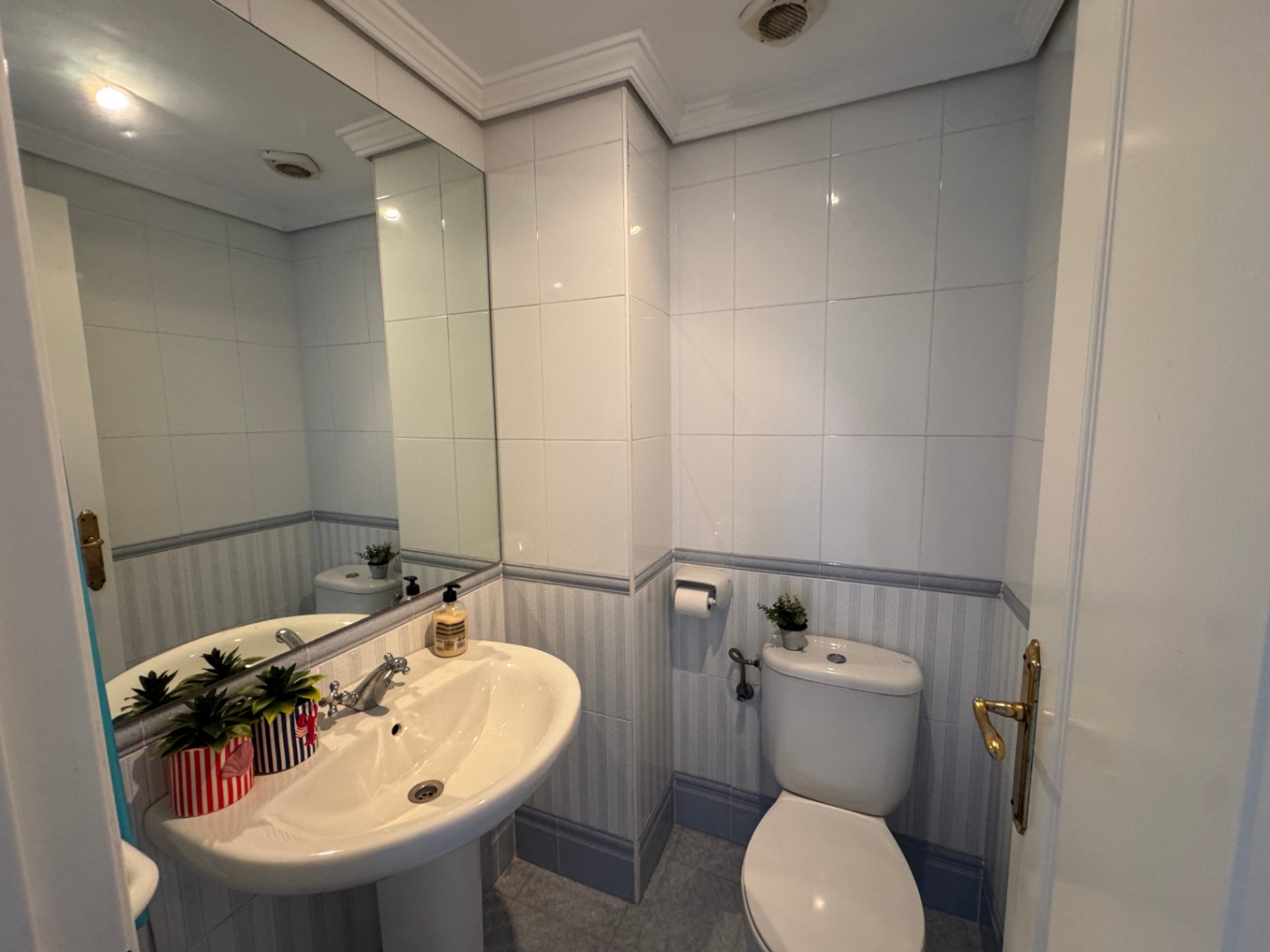 Functional bathroom featuring a white pedestal sink, large frameless mirror, and tiled walls with a blue-striped decorative border.