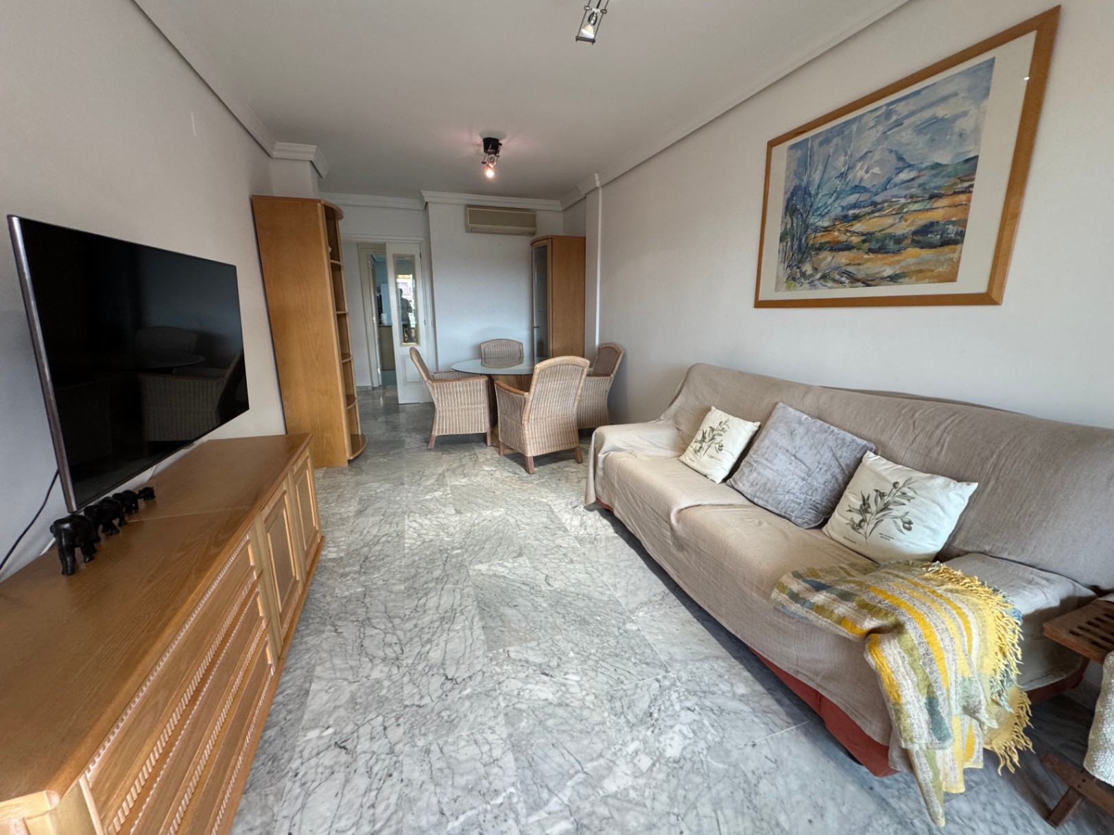 Living area featuring grey marble flooring, a wooden TV console, and a dining space with a glass-top table and four wicker chairs.