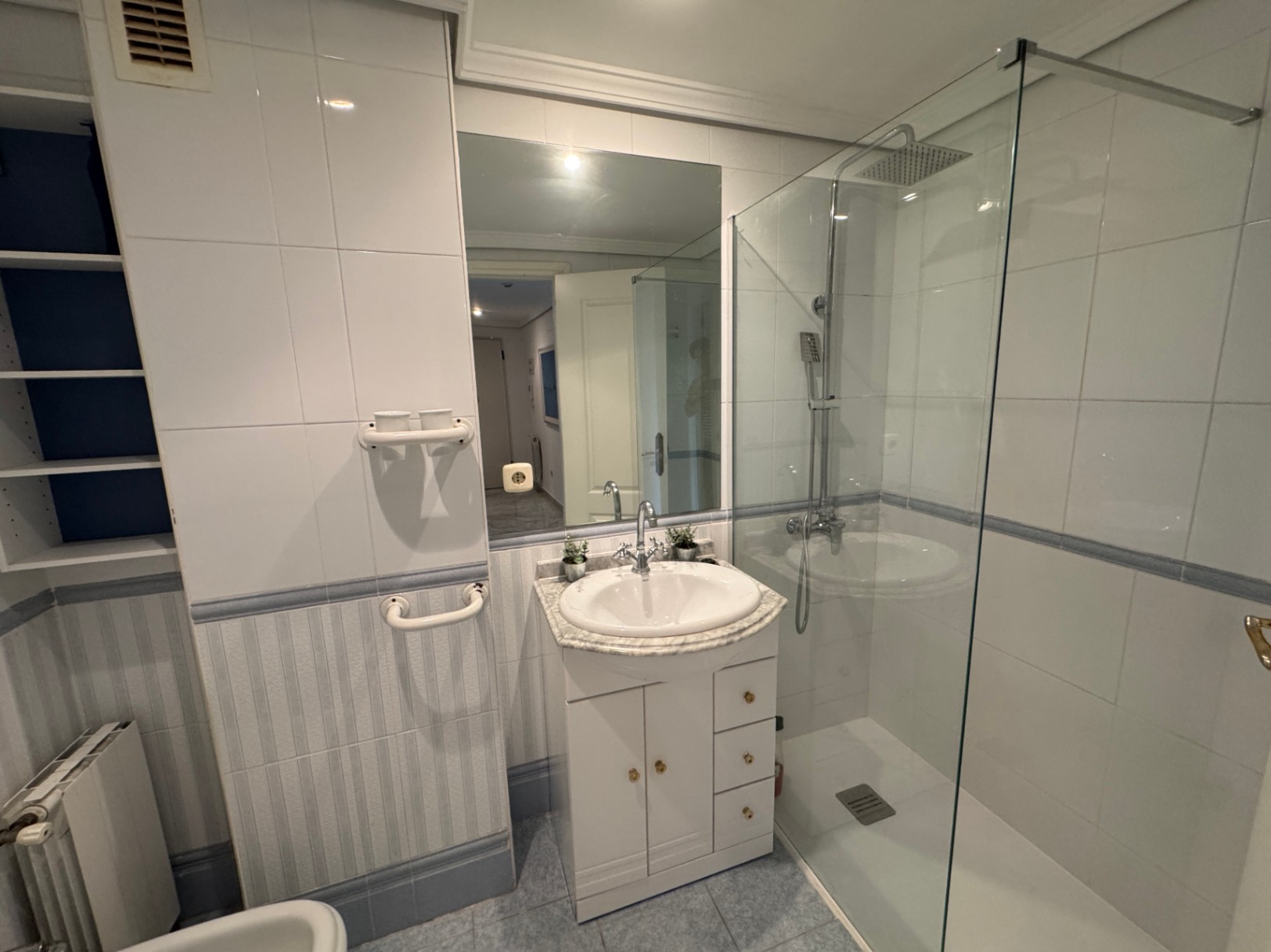 Modern bathroom featuring a walk-in glass shower, a white vanity with marble countertop, and integrated shelving. The walls are finished with white and striped ceramic tiles.