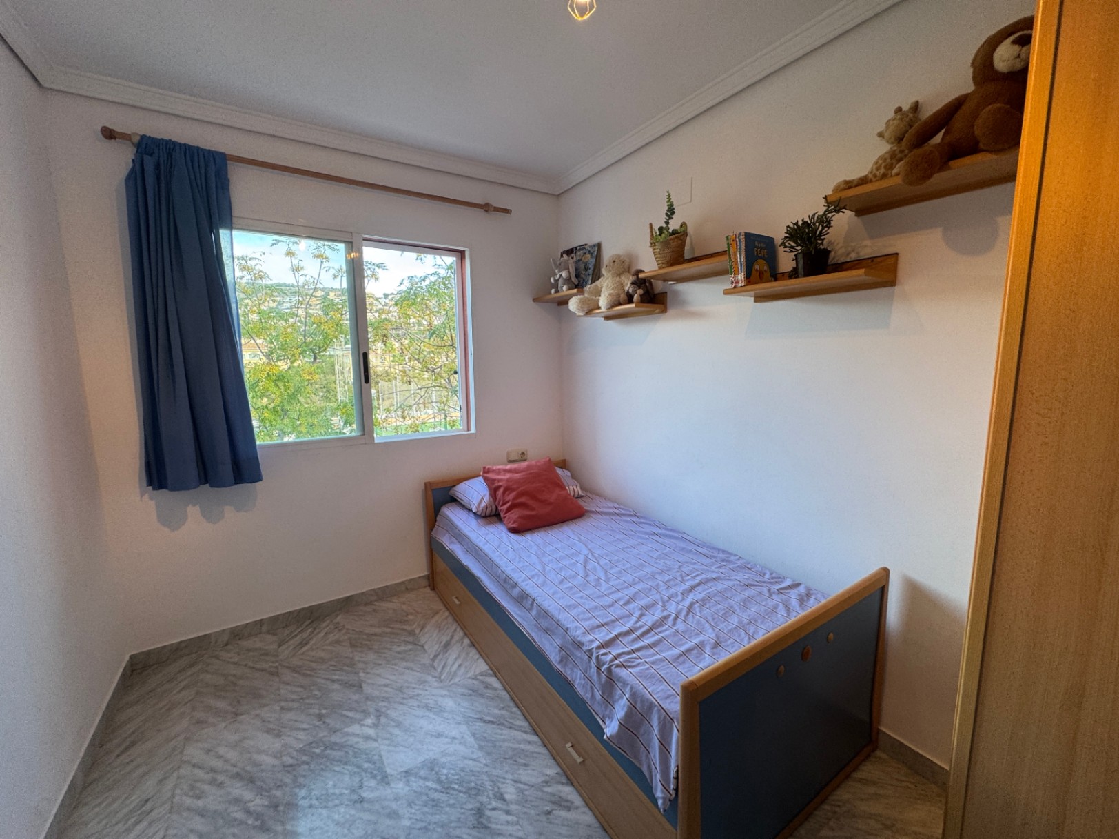 Single bedroom featuring marble flooring, wooden floating shelves, and a sliding window with garden views. The bed frame includes integrated storage drawers.