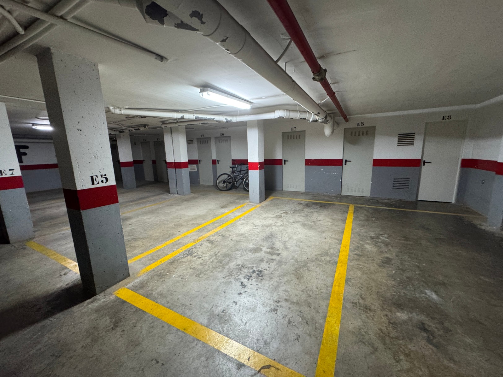 Underground parking garage with concrete flooring, yellow markings, and numbered storage doors. Features exposed overhead pipes and fluorescent lighting.