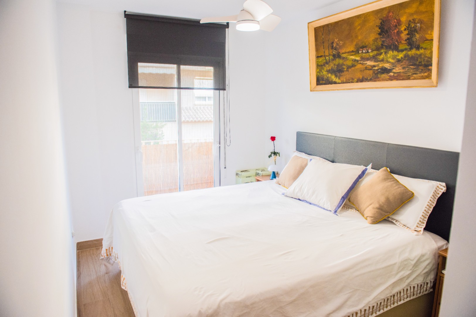 Bright bedroom featuring a grey headboard, ceiling fan, and large window with a black roller blind. A landscape oil painting hangs above the bed.