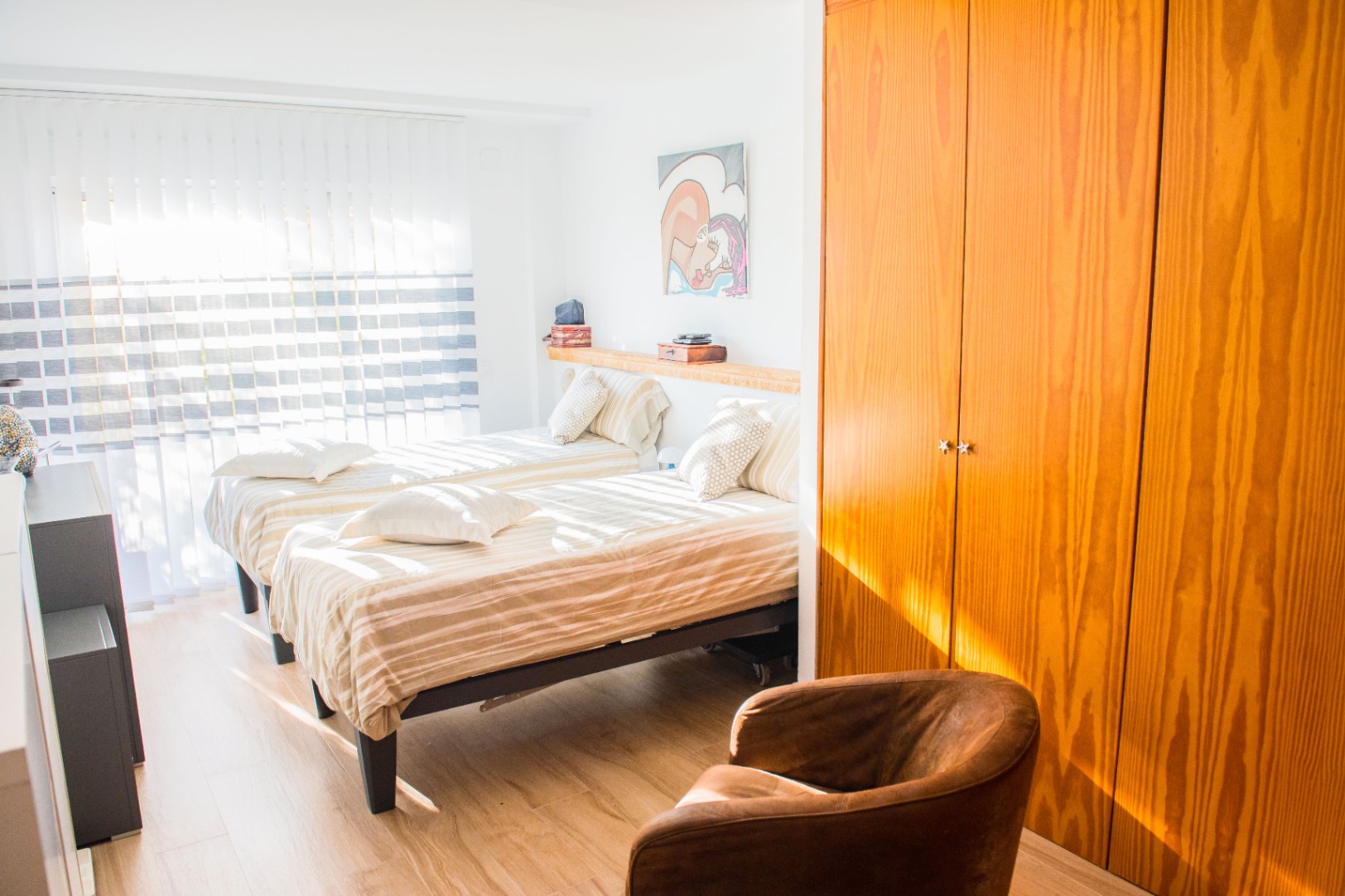 Bright bedroom featuring twin beds with striped linens, a large built-in wooden wardrobe, and a leather armchair on light wood flooring.