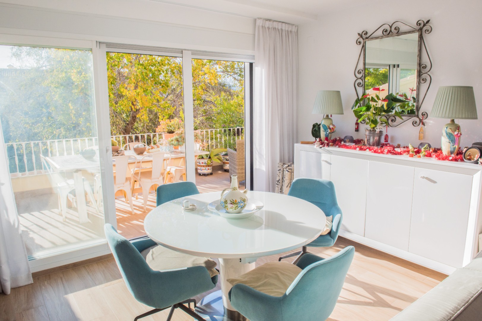 Bright dining area featuring a round white table and blue chairs, with direct access to a sunlit terrace through large sliding glass doors.