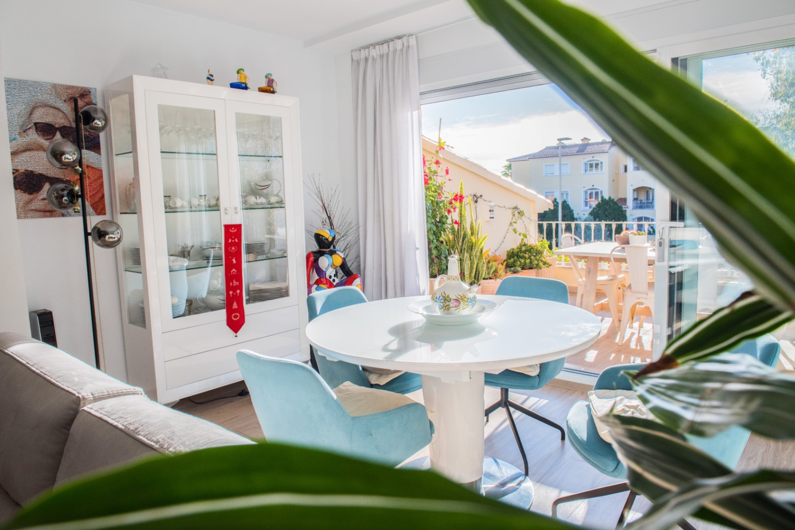 Bright dining area featuring a round white table, blue upholstered chairs, and a white display cabinet, with direct access to a sunlit terrace.