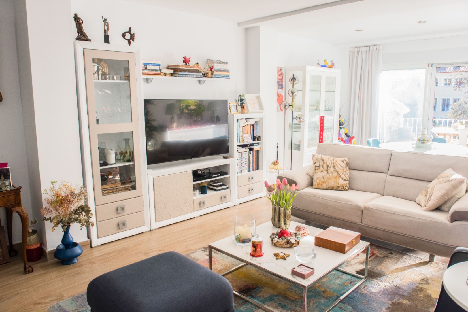Bright living area featuring a beige fabric sofa, marble-top coffee table, and a white modular media unit. Large glass doors provide ample natural light to the room.