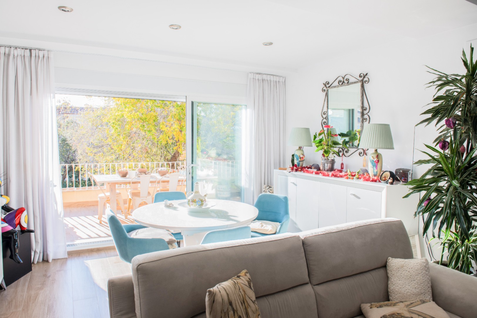 Bright living area featuring a round white dining table with blue chairs, adjacent to a terrace with outdoor seating and views of lush greenery.
