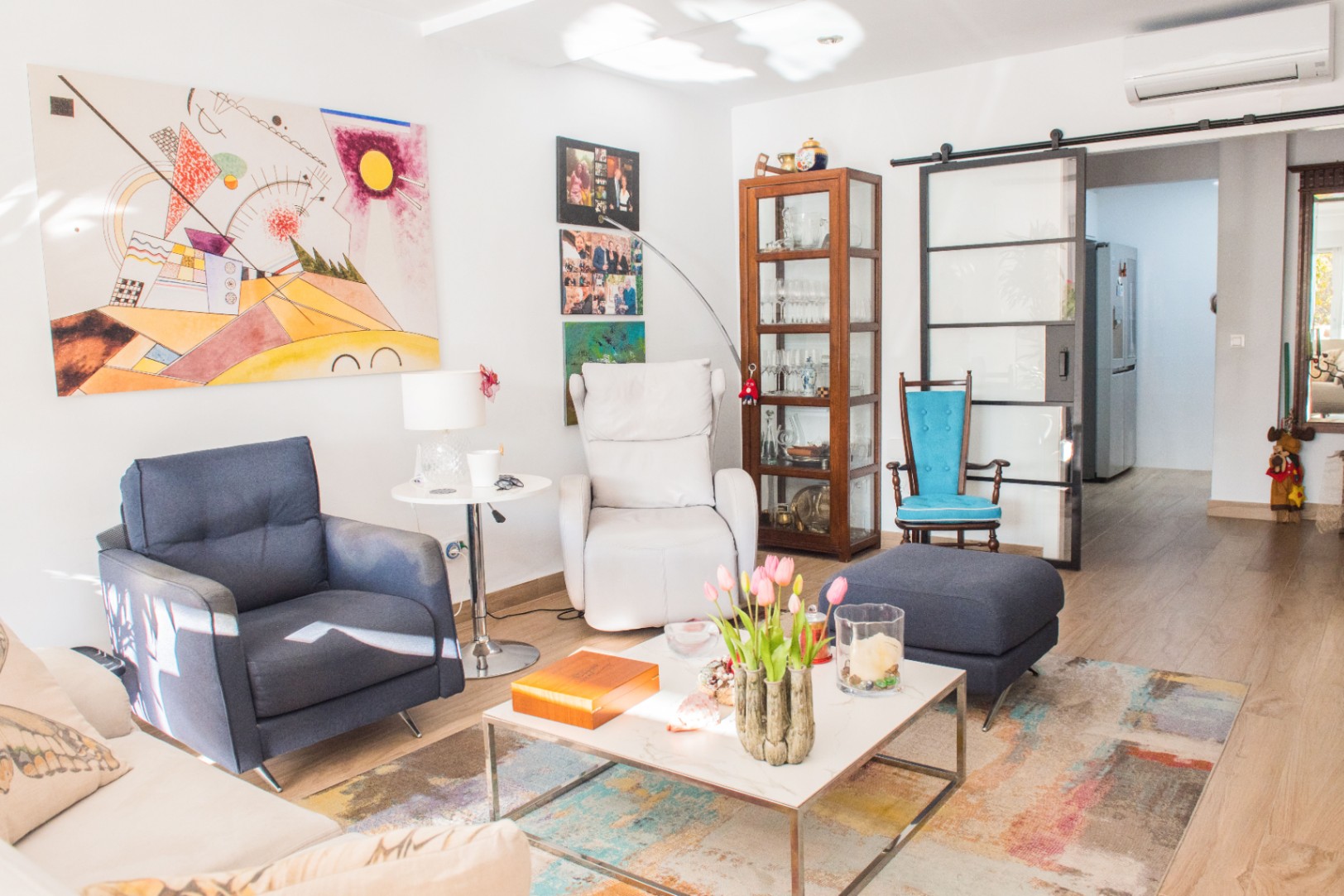 Bright living room featuring a blue armchair, white recliner, and abstract wall art. A black-framed sliding glass door separates the space from the hallway.
