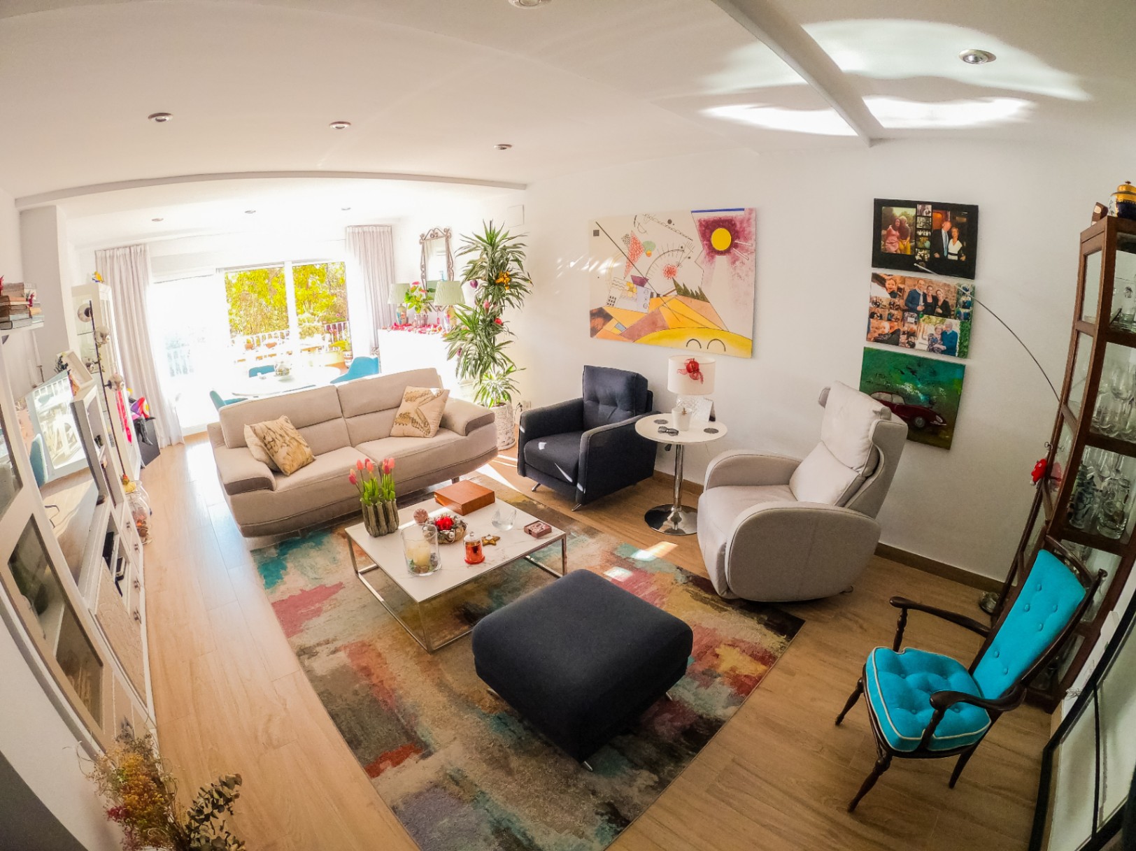 Bright living room featuring light wood flooring, a multi-colored abstract rug, and varied seating. Large glass doors provide natural light and access to a terrace with greenery.