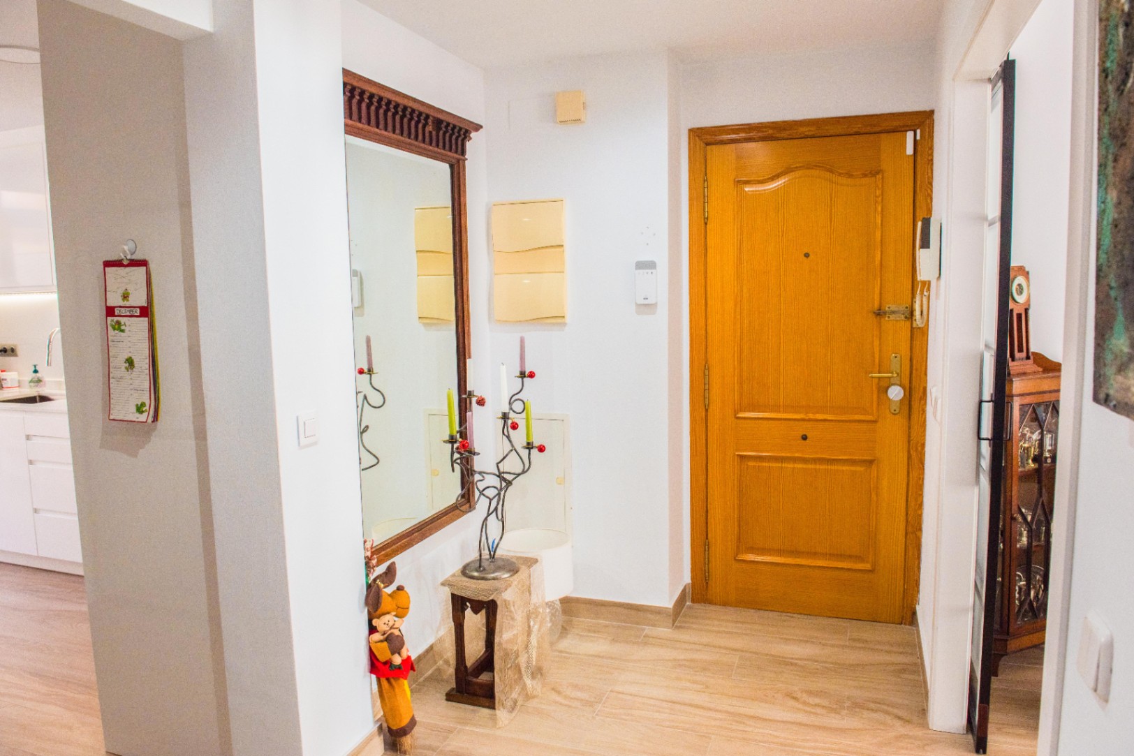 Entrance hall featuring a solid wood door, light-toned tiled flooring, and a large mirror with a dark wood frame. The space connects to a bright kitchen.