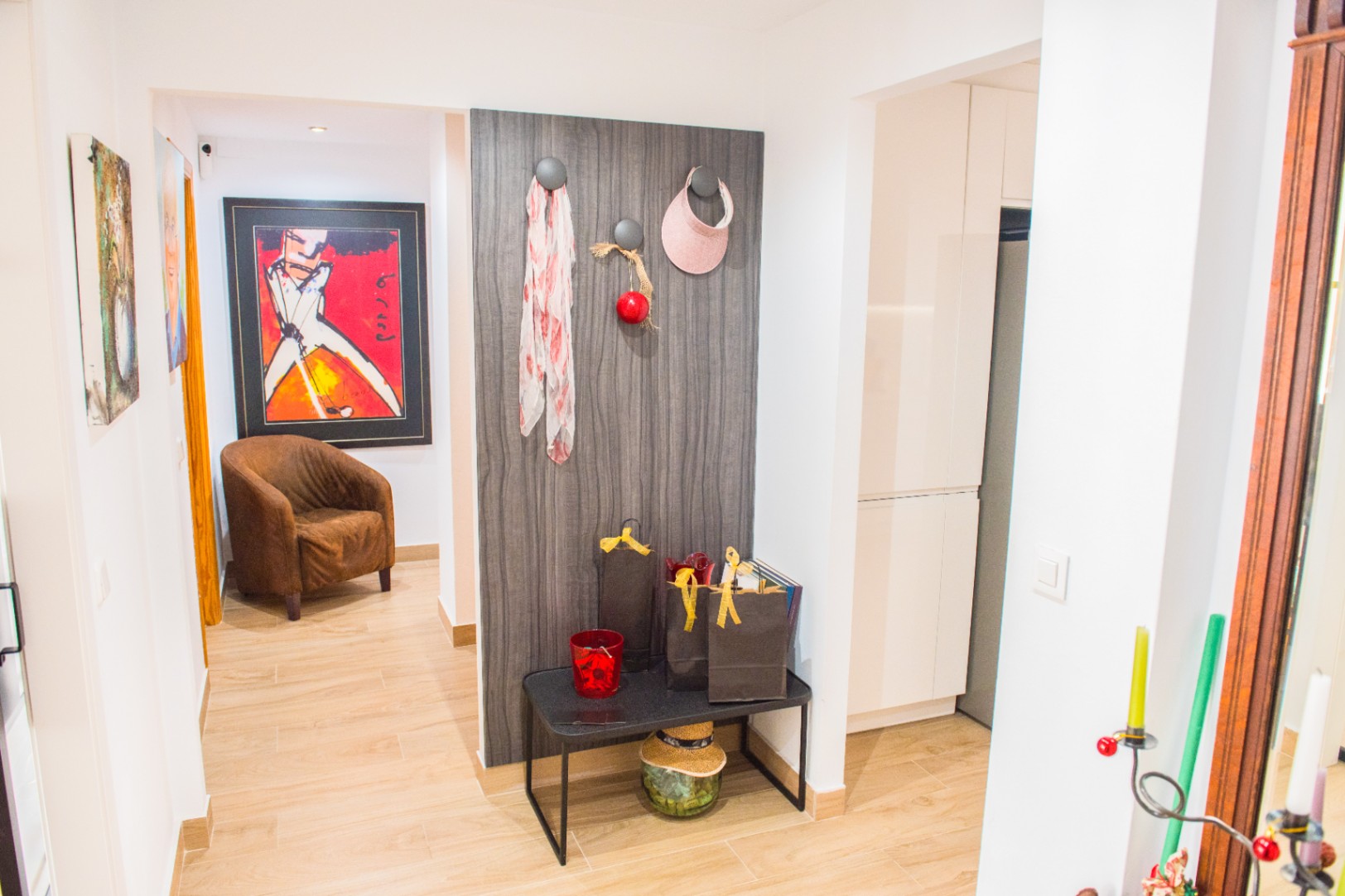 Entryway featuring wood-effect flooring, a dark timber-grain accent panel with hooks, and a minimalist black bench. A vibrant art piece and brown armchair define the background hallway.