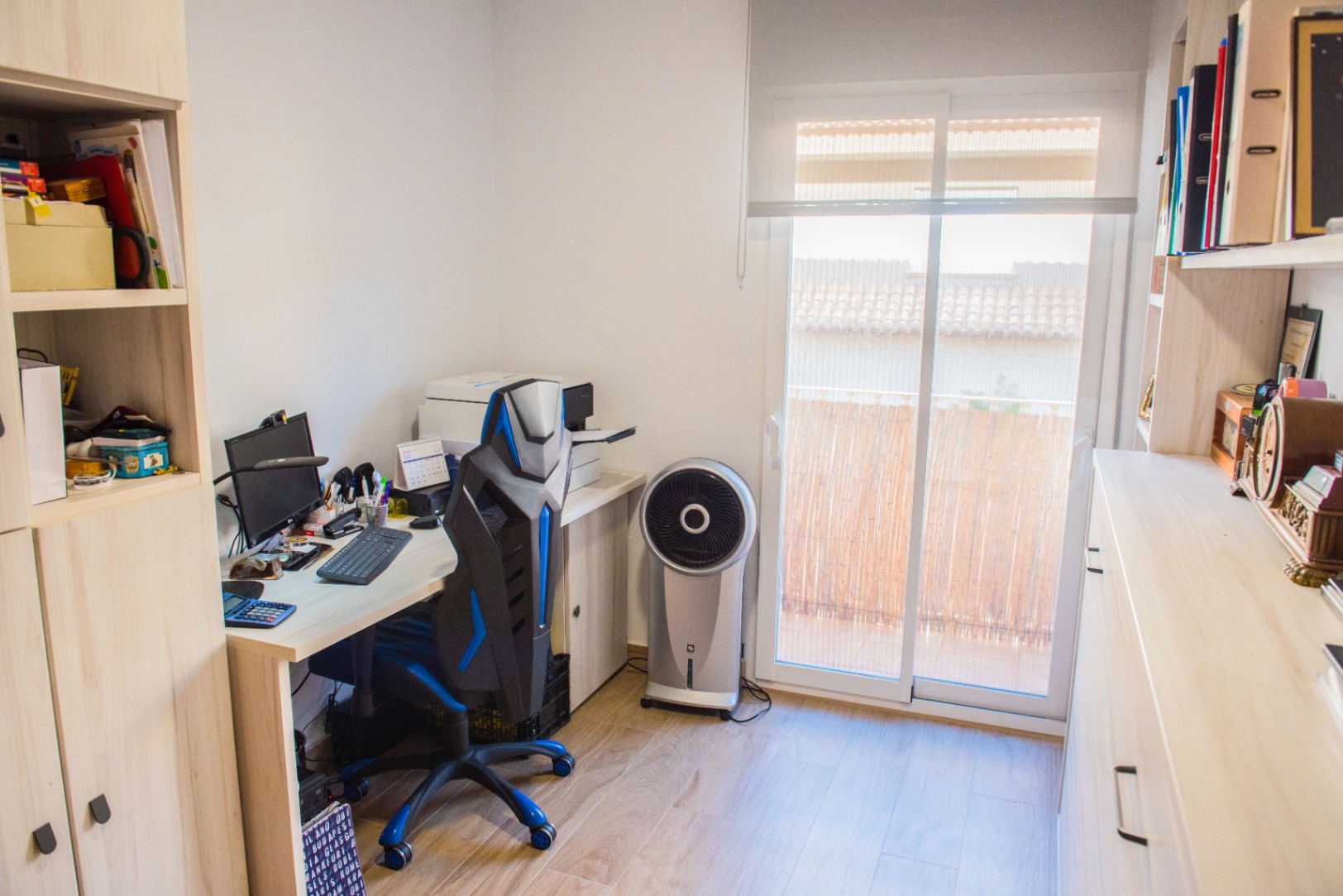 Home office with a light wood desk, ergonomic chair, and integrated shelving. A sliding glass door provides natural light and access to a balcony with a bamboo screen.