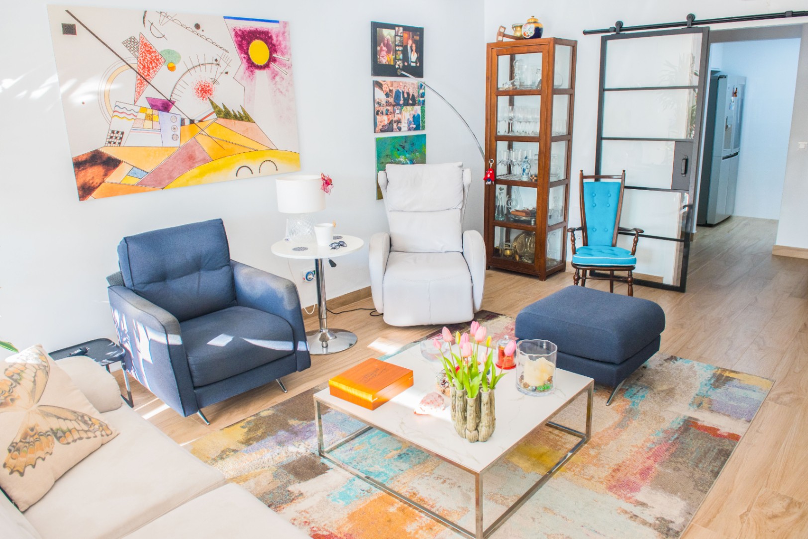 Living area featuring a blue armchair, white recliner, and abstract art. Includes a wooden display cabinet and a black-framed sliding glass door over wood-effect flooring.