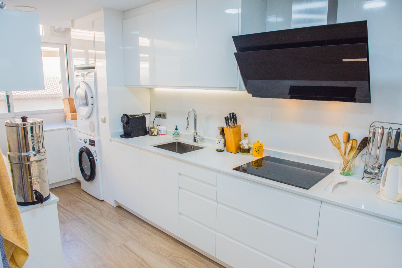 Minimalist white kitchen featuring high-gloss cabinetry, an induction cooktop, and an integrated laundry nook with a stacked washer and dryer.