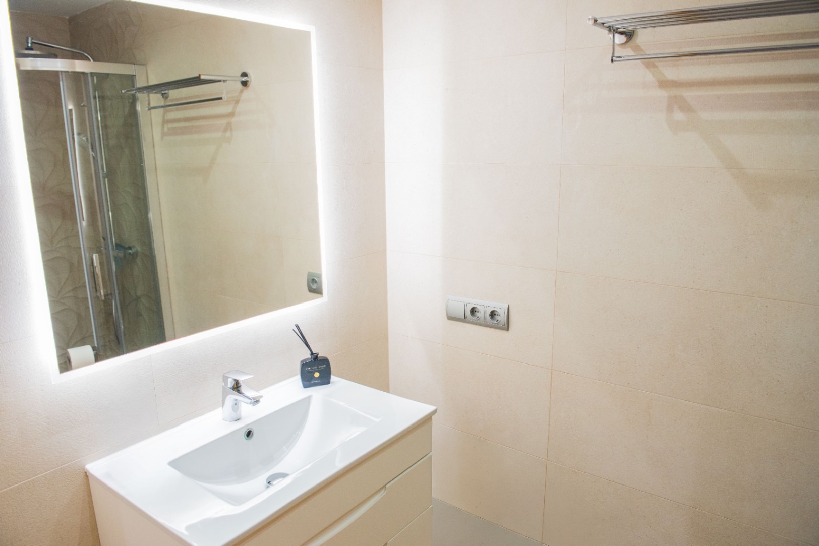 Modern bathroom featuring a white vanity sink, a large mirror with integrated LED lighting, and neutral cream-toned wall tiling.