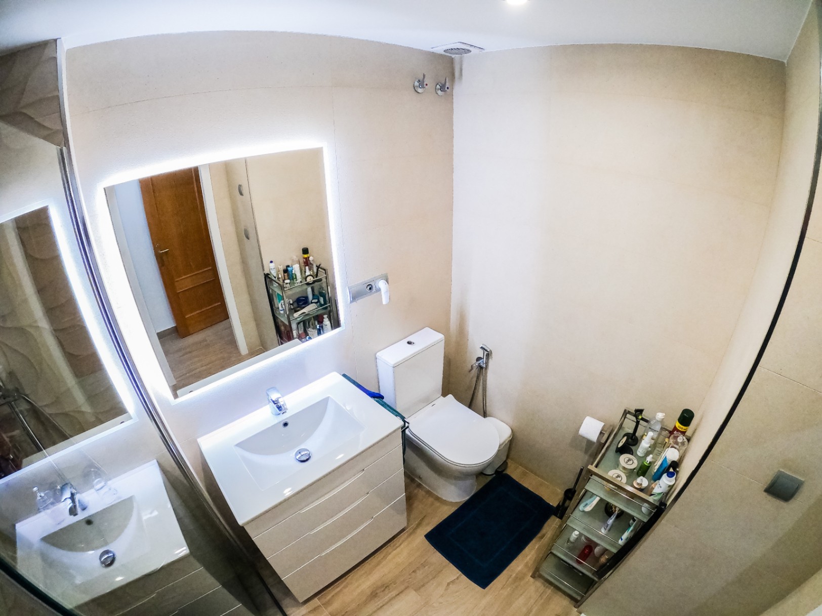 Modern bathroom featuring a white vanity with integrated sink, a backlit LED mirror, and wood-effect flooring. The space includes a floor-mounted toilet and beige tiled walls.