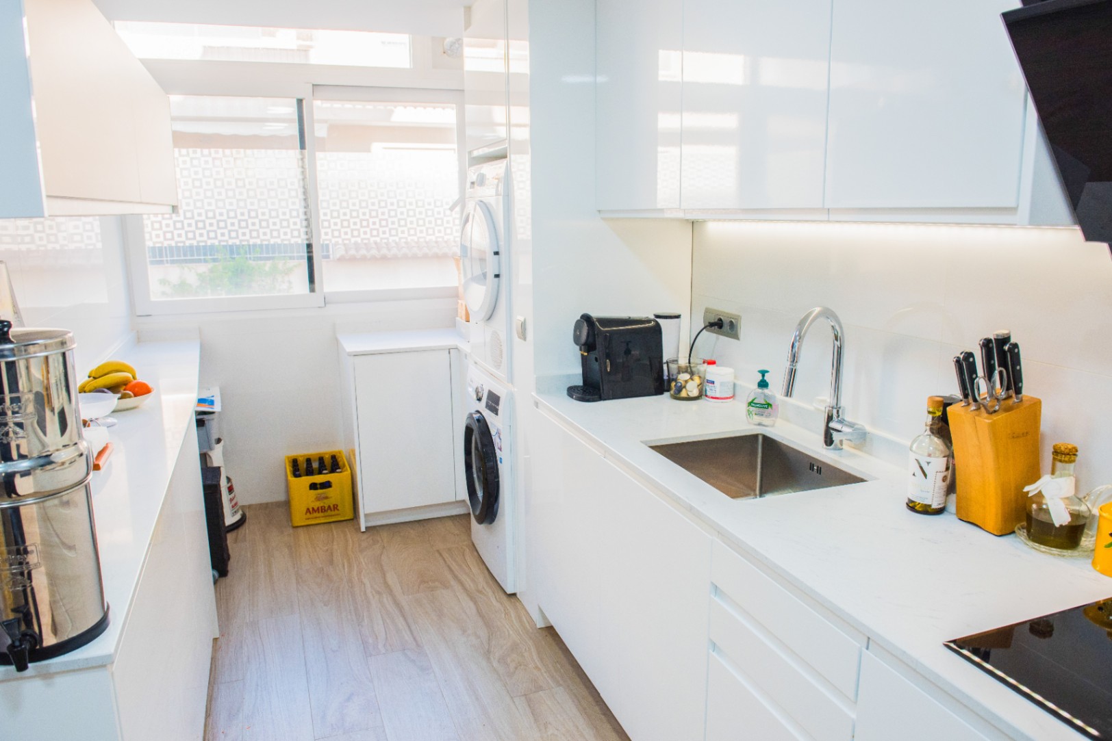 Modern galley kitchen with white handleless cabinetry, integrated laundry appliances, and under-cabinet LED lighting. Features include a stainless steel sink and light wood flooring.