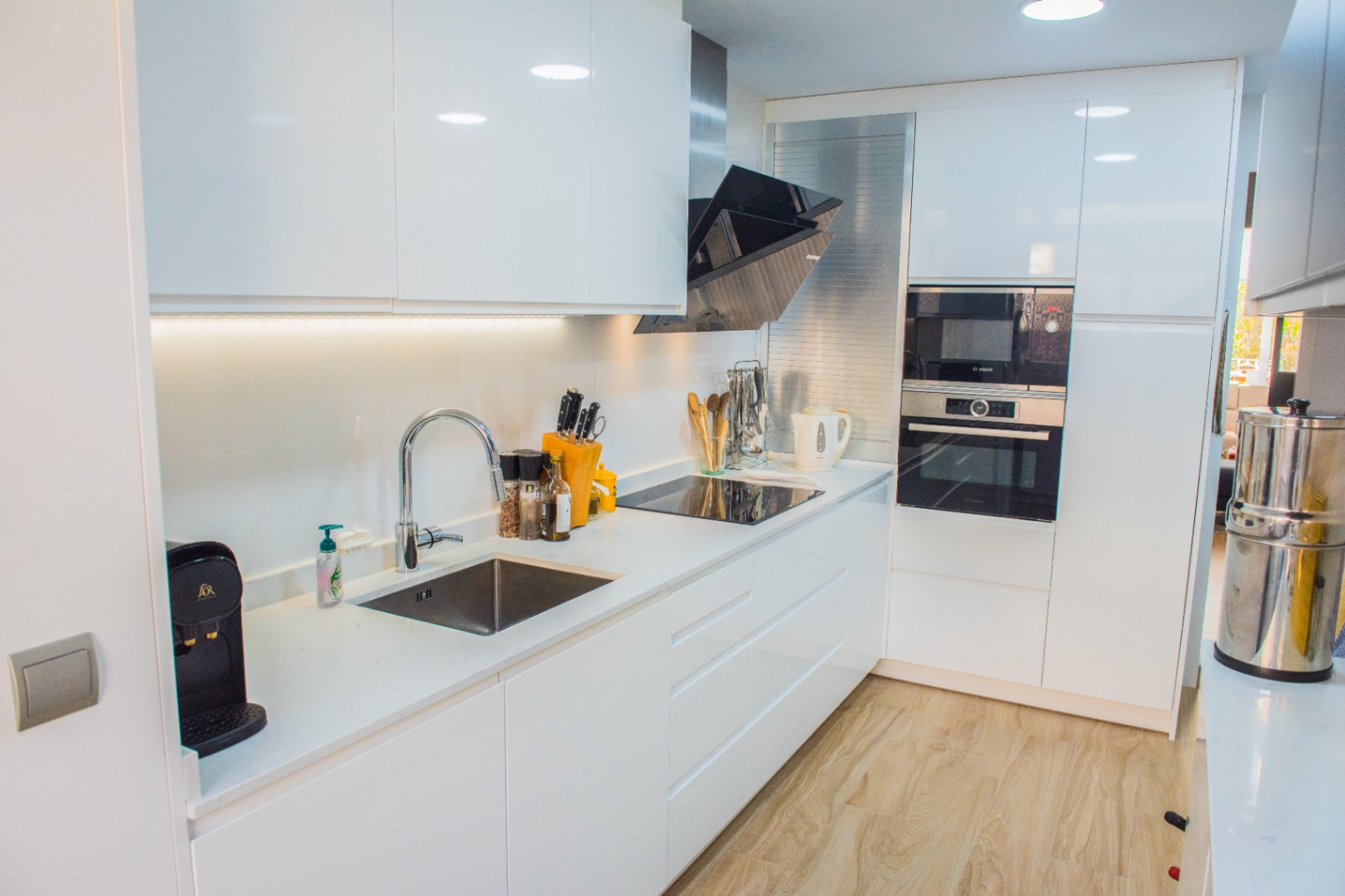 Modern kitchen with white high-gloss cabinetry, integrated Bosch appliances, and an induction cooktop. Features a white quartz countertop and light wood-effect flooring.