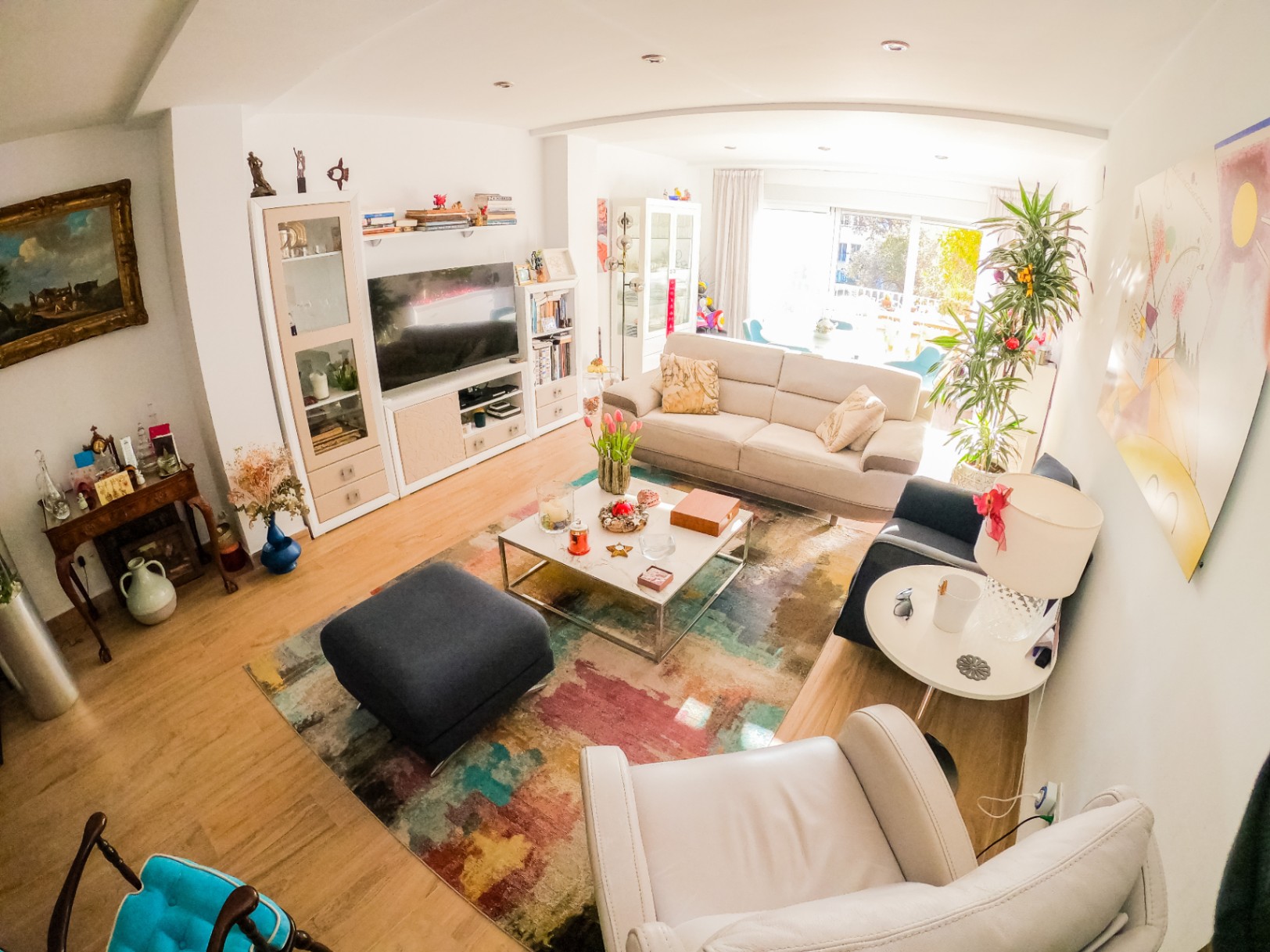 Spacious living room featuring a colorful abstract rug, light wood flooring, and a beige sofa. Large sliding glass doors provide abundant natural light and access to a balcony.