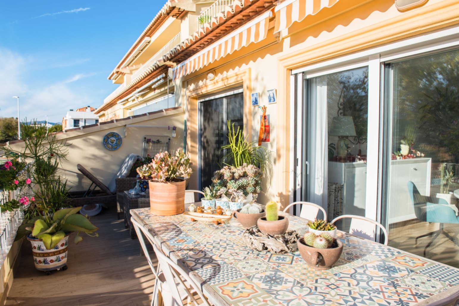 Sunny Mediterranean terrace featuring a tiled mosaic dining table, potted succulents, and large sliding glass doors under a striped retractable awning.