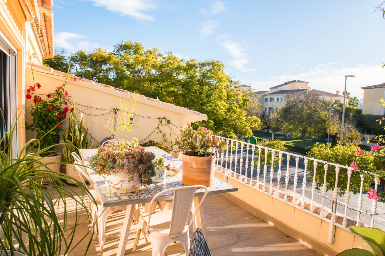 Terrace featuring a tiled mosaic dining table, white metal chairs, and potted succulents overlooking a residential area with mature trees.