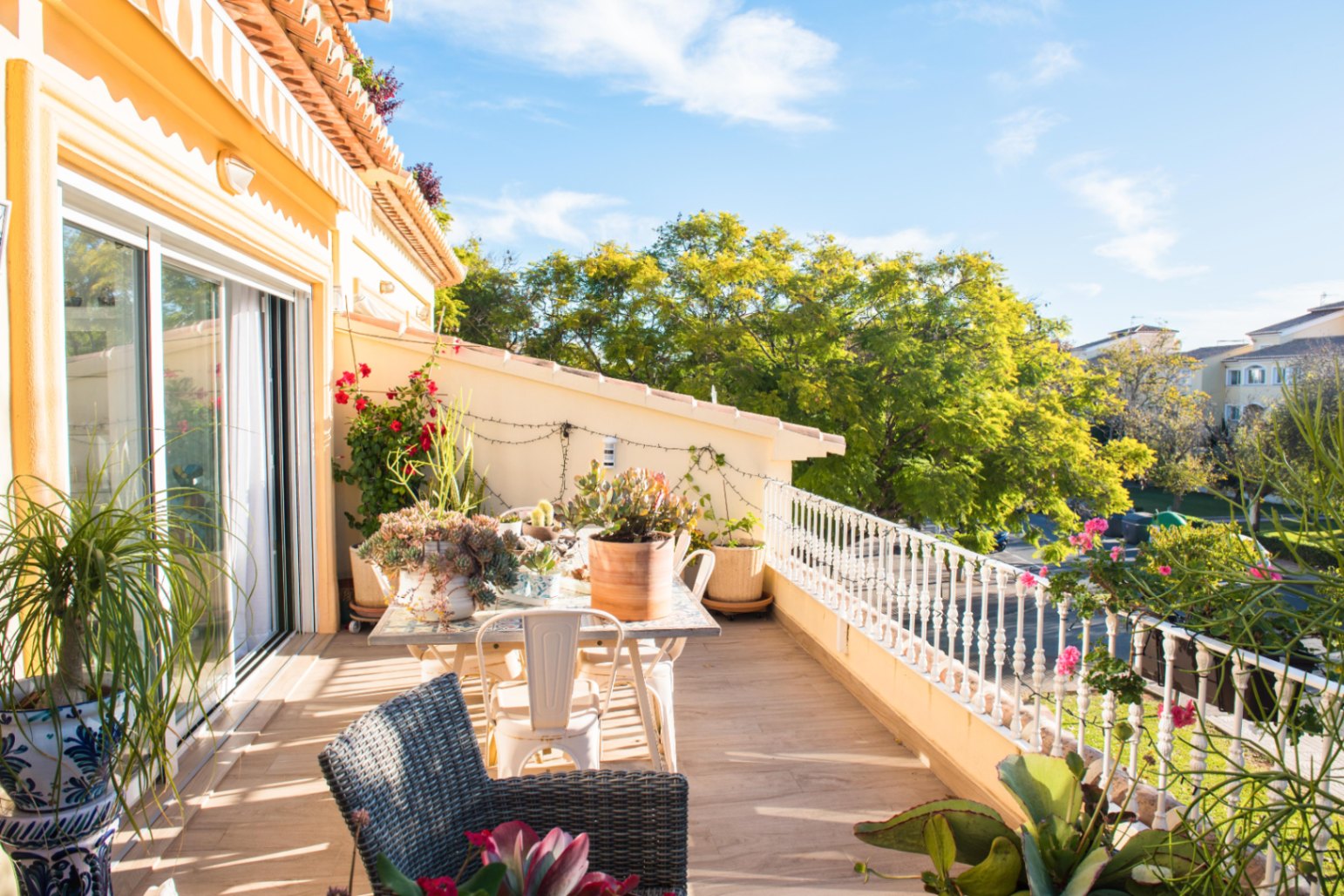 Terrace with wood-effect tiling, outdoor dining area, and white balustrade. Surrounded by potted plants and views of lush greenery under a clear sky.