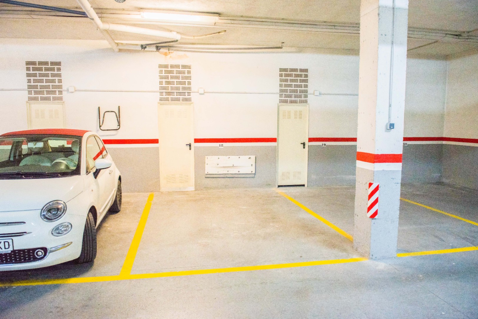 Underground parking space featuring numbered bays, yellow floor markings, and white walls with a red accent stripe and exposed overhead pipes.