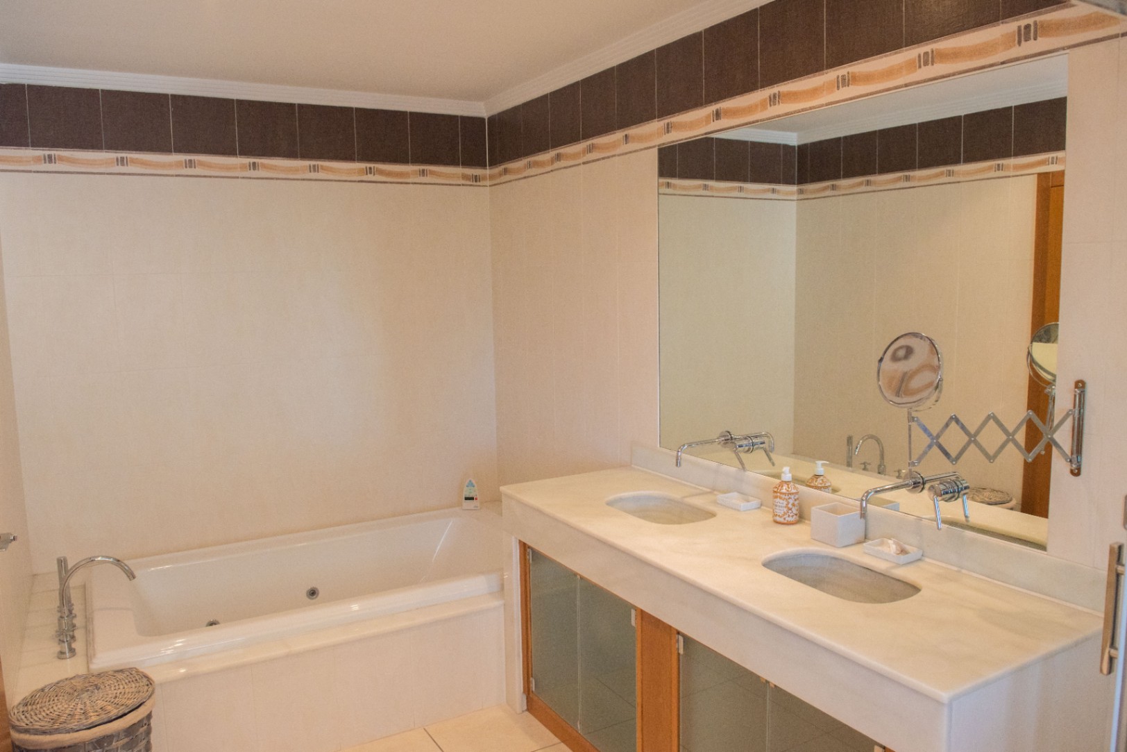 Bathroom featuring a double vanity with stone countertops, wall-mounted faucets, and a built-in jetted bathtub set against tiled walls with a decorative border.
