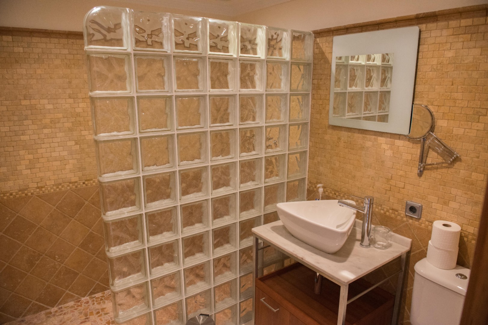Bathroom featuring a glass block shower partition, tan mosaic wall tiles, and a white vessel sink on a marble countertop with wooden storage.
