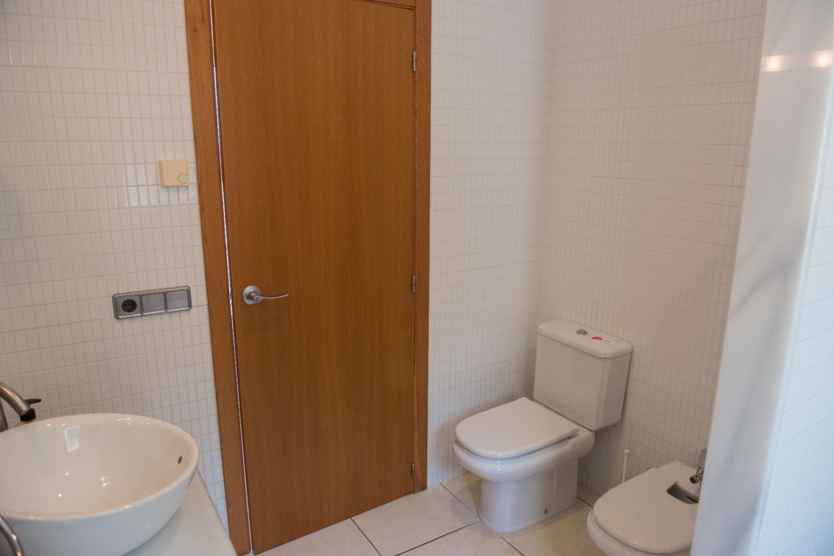 Bathroom with white square tiles, a wooden door, ceramic toilet, bidet, and a white vessel sink with chrome fittings.