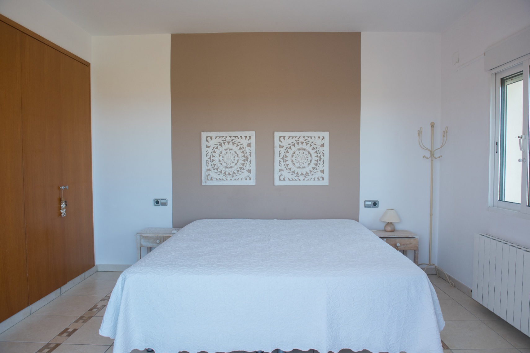 Bedroom featuring a taupe accent wall with carved white panels, a double bed, integrated wooden wardrobe, and tiled flooring with decorative borders.