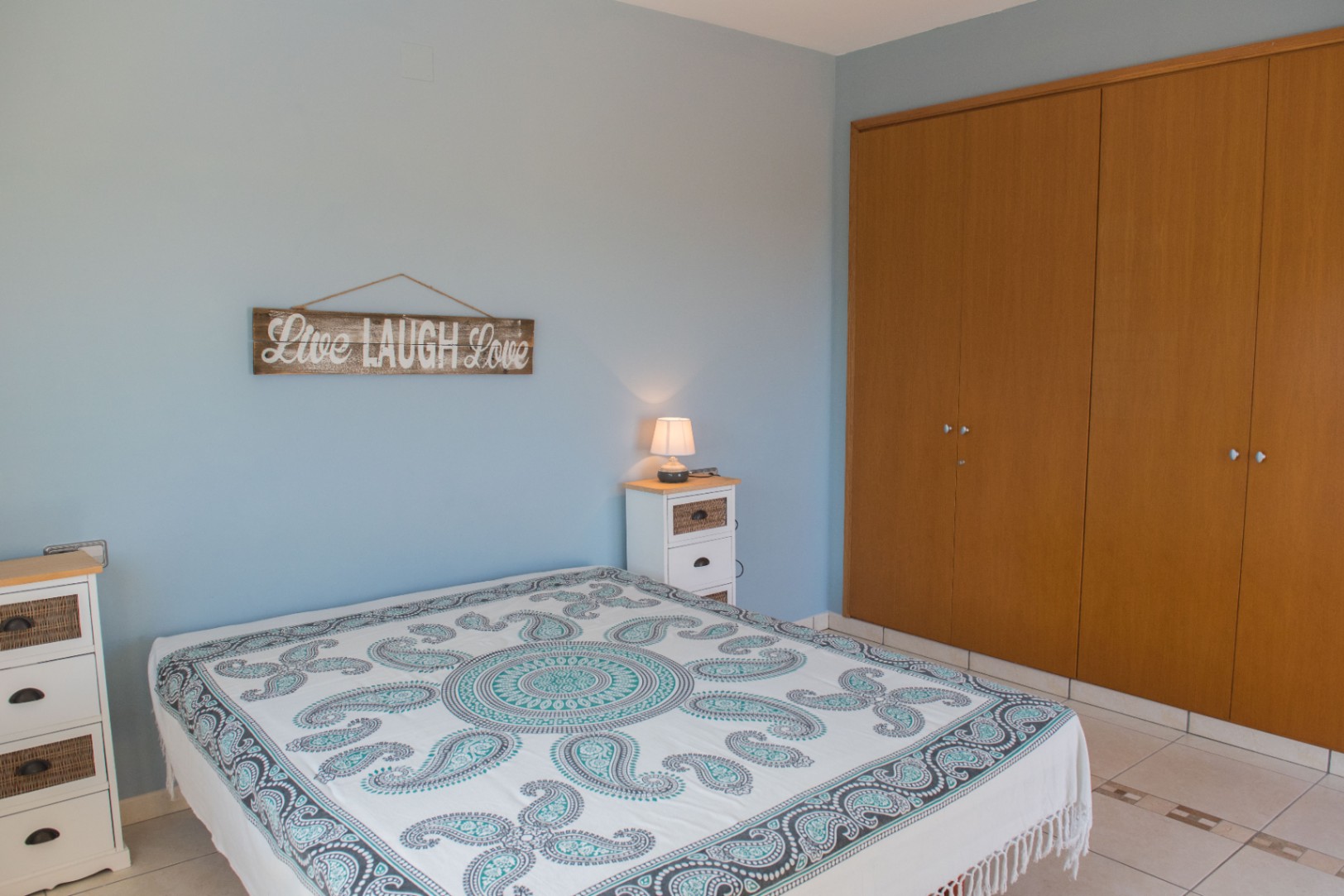 Bedroom featuring light blue walls, a large built-in wooden wardrobe, and white furniture with wicker details. A patterned bedspread and tiled flooring complete the space.