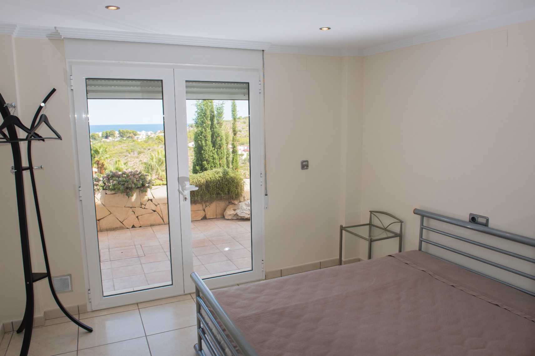 Bedroom with a grey metal bed frame and glass side table, featuring double doors that open onto a tiled terrace with coastal views.
