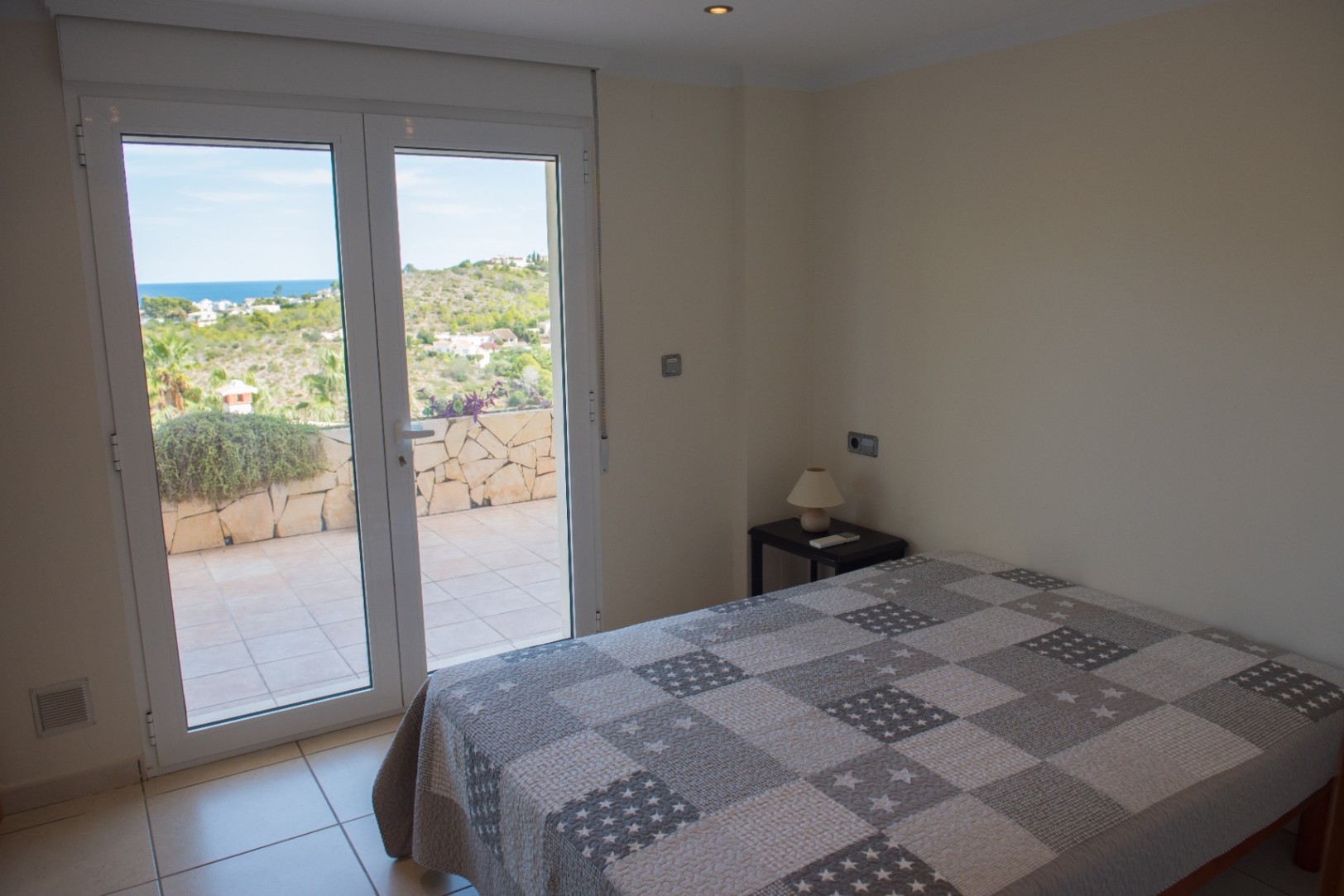 Bedroom with direct access to a tiled terrace through glass doors, offering views of the Mediterranean landscape and sea. Features neutral-toned decor and tile flooring.
