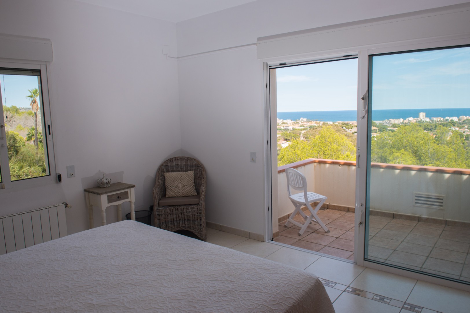 Bedroom with private balcony access offering panoramic sea and coastal views. Features white tiled flooring, a wicker armchair, and a large sliding glass door.