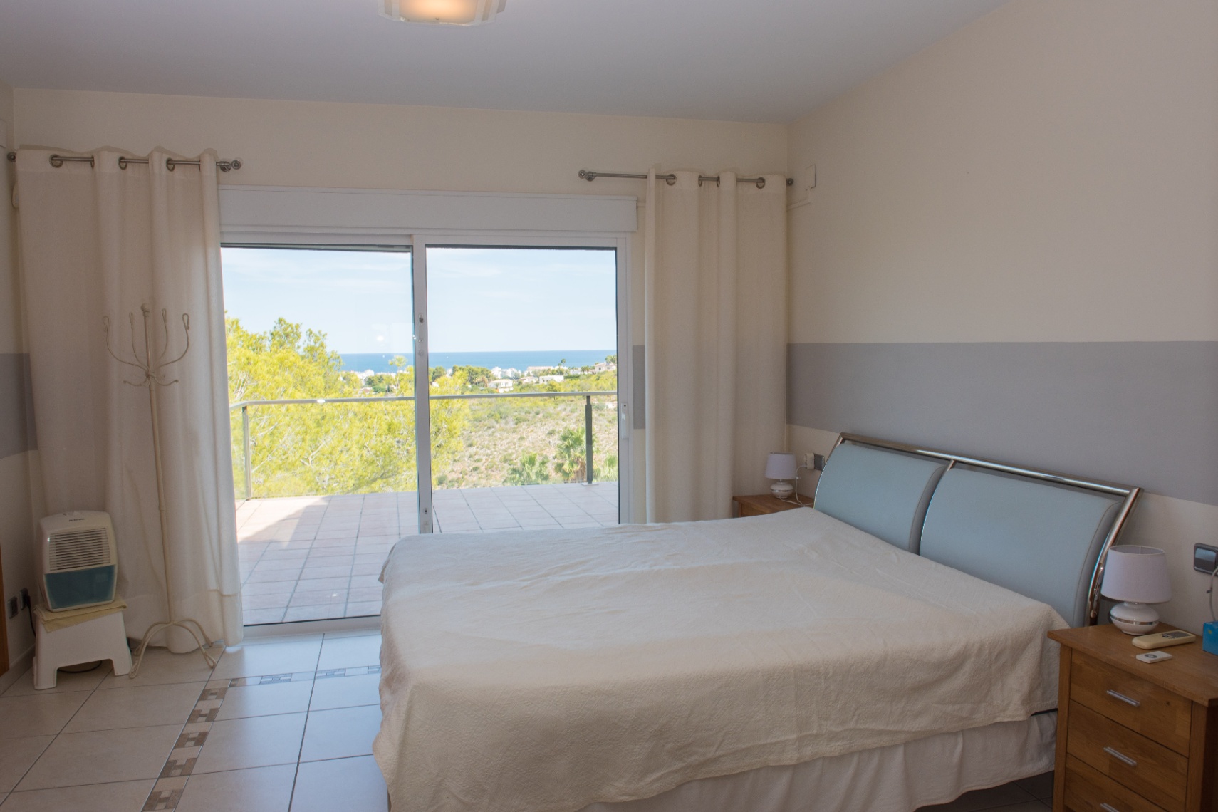 Bedroom with tiled flooring and sliding glass doors opening to a terrace with sea views. Features a modern metallic headboard and neutral two-tone walls.