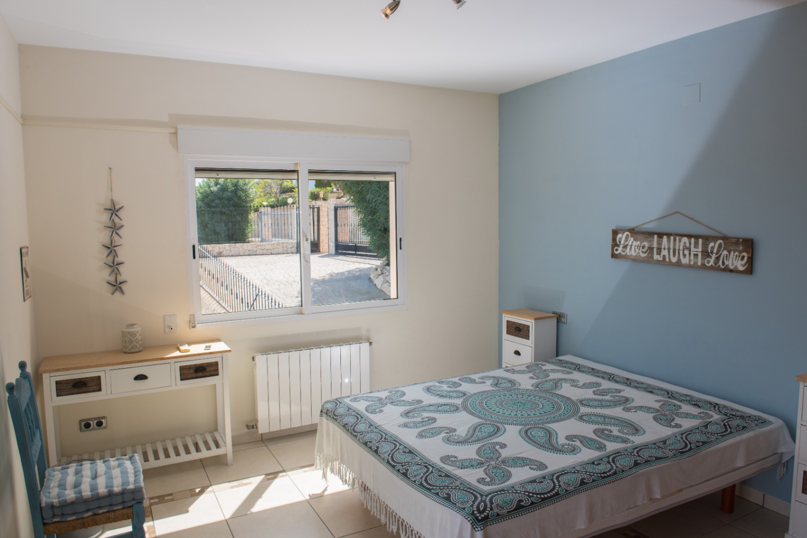 Bright bedroom featuring a blue accent wall, tiled flooring, and a large window overlooking the private gated courtyard. Includes a white console table and radiator.