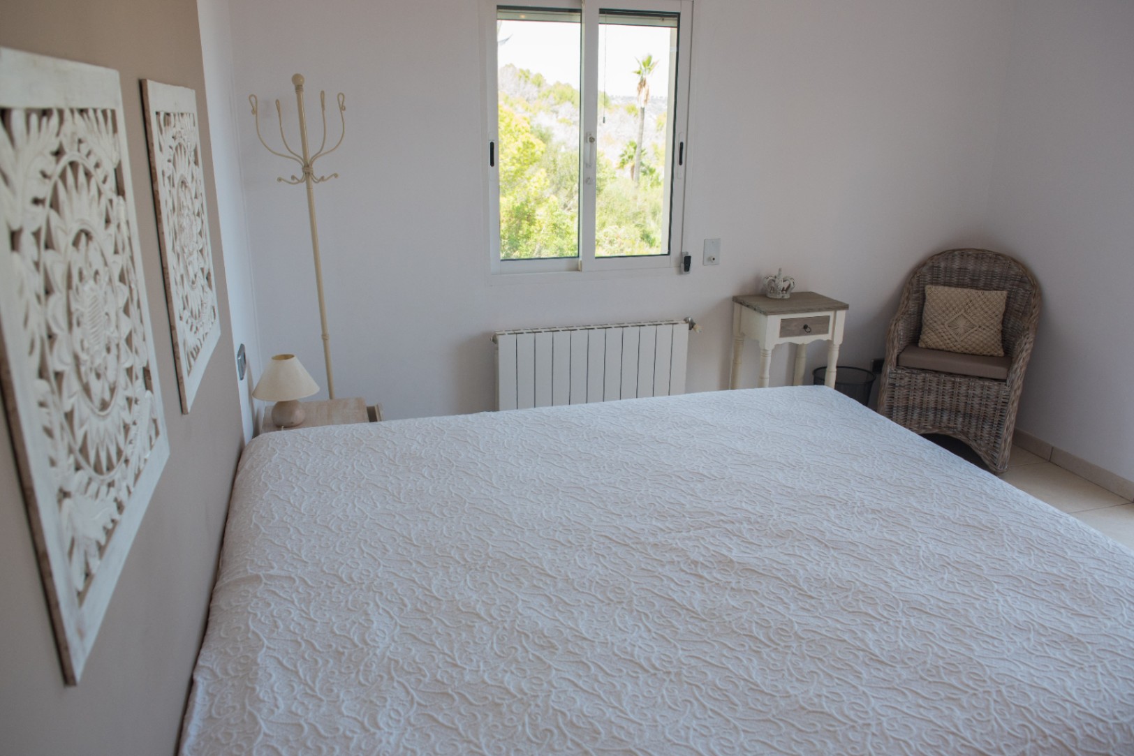 Bright bedroom featuring a textured white bedspread, carved wall art, and a wicker armchair. A window offers views of Mediterranean greenery and palm trees.