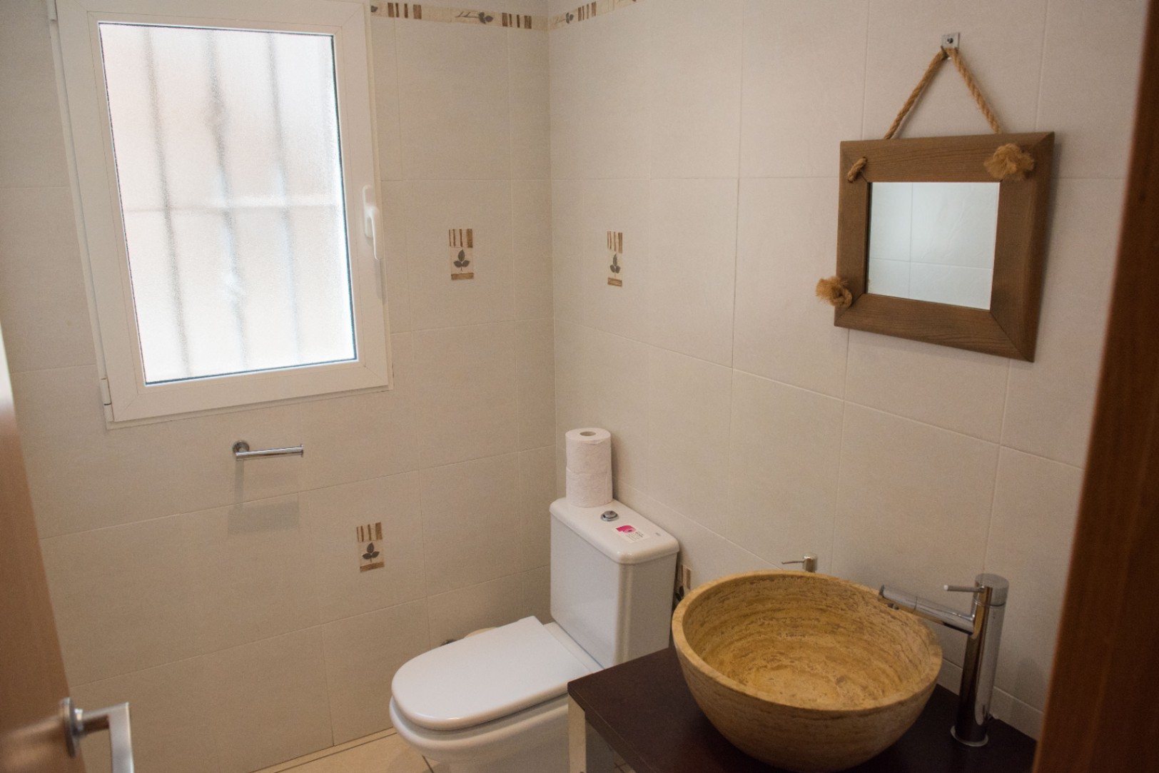 Compact bathroom featuring a natural stone vessel sink on a dark wood vanity, complemented by a rustic rope-hung mirror and neutral tiled walls.