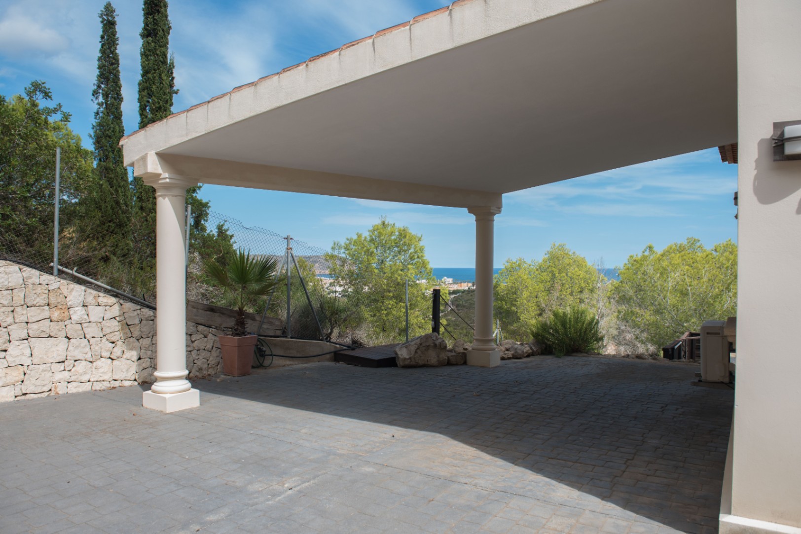Covered parking area featuring classical white columns and grey stone paving, offering views of the Mediterranean coastline and surrounding greenery.