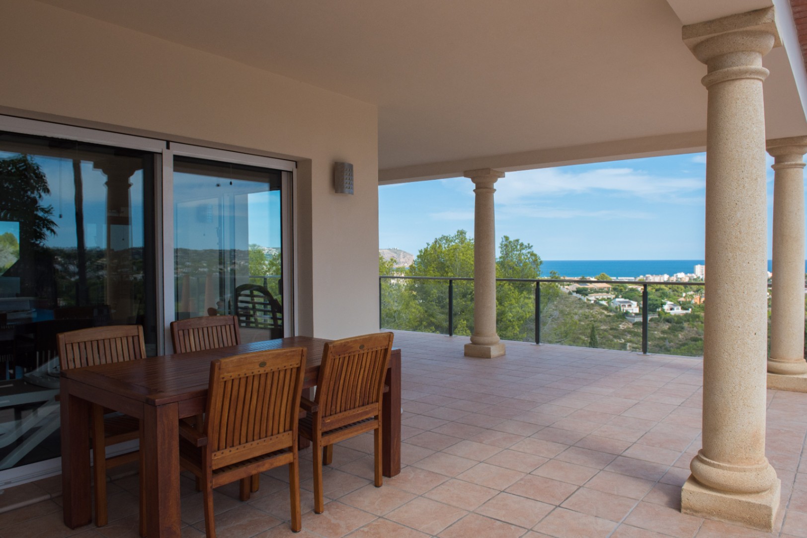 Covered terrace featuring a wooden dining set, classic stone columns, and tiled flooring. Offers elevated coastal views through a glass balustrade.
