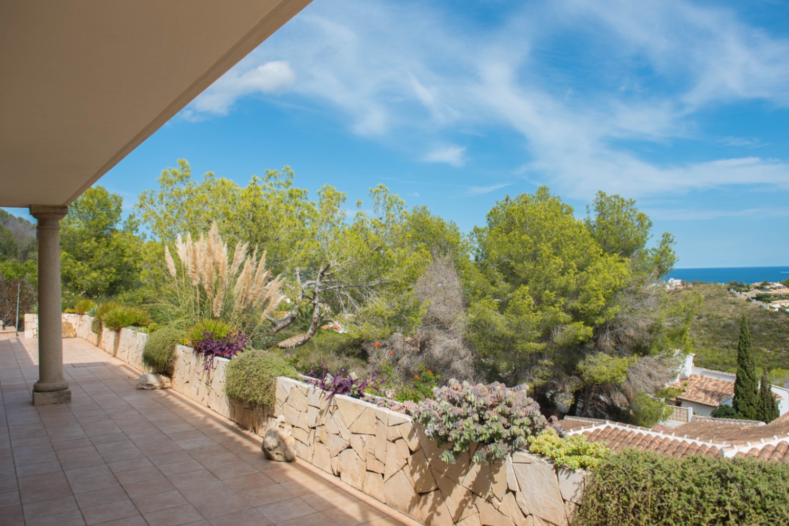 Covered terrace with terracotta tiling and a stone-clad planter featuring Mediterranean flora, offering views of pine trees and the distant sea.