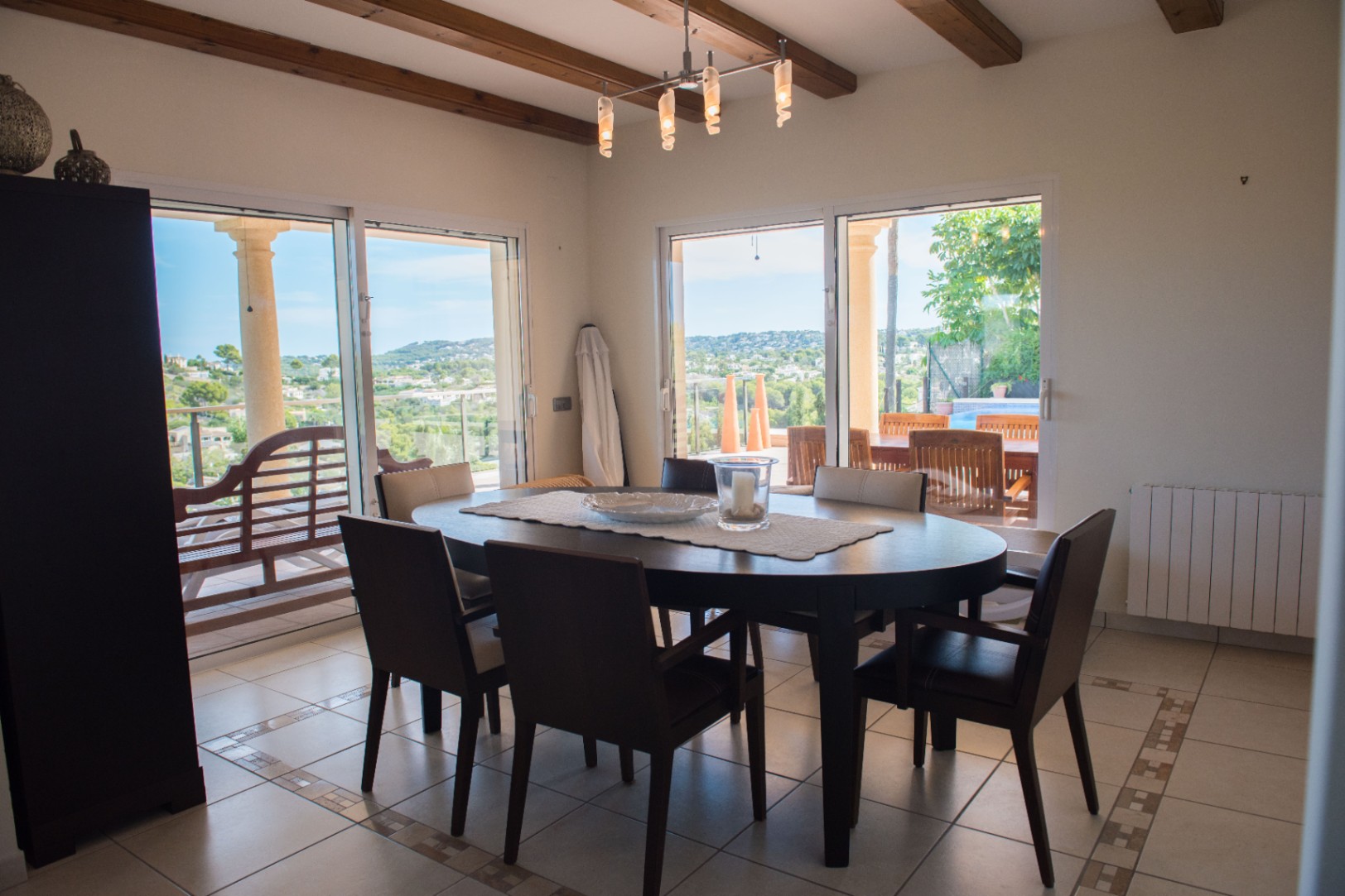 Dining area featuring an oval dark wood table and exposed ceiling beams. Large sliding glass doors offer panoramic views of the valley and direct access to the terrace.