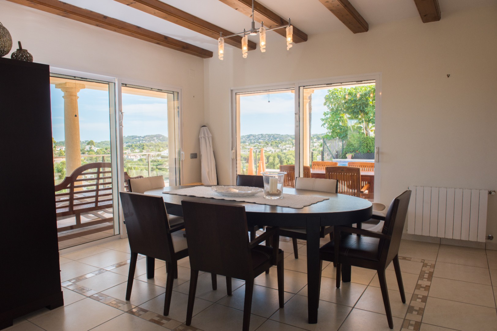 Dining area with an oval dark wood table, exposed ceiling beams, and large sliding glass doors providing direct access to the terrace and panoramic hillside views.
