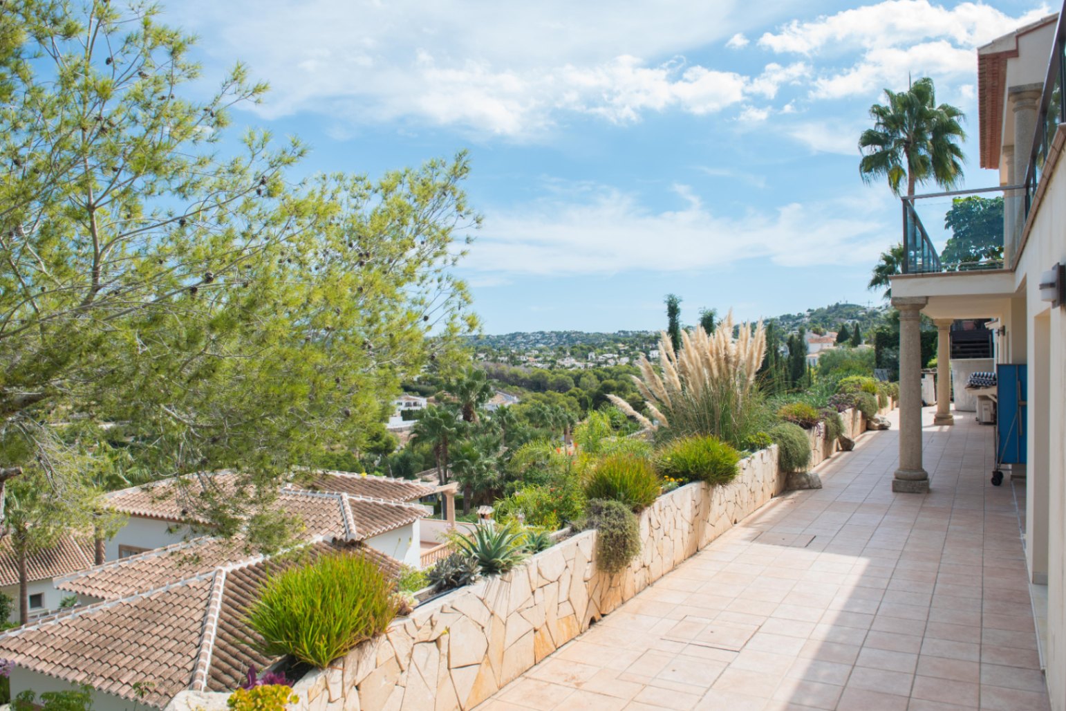 Elevated terrace with beige tiles and a stone planter wall featuring Mediterranean vegetation. Offers panoramic views of the valley and surrounding villas.